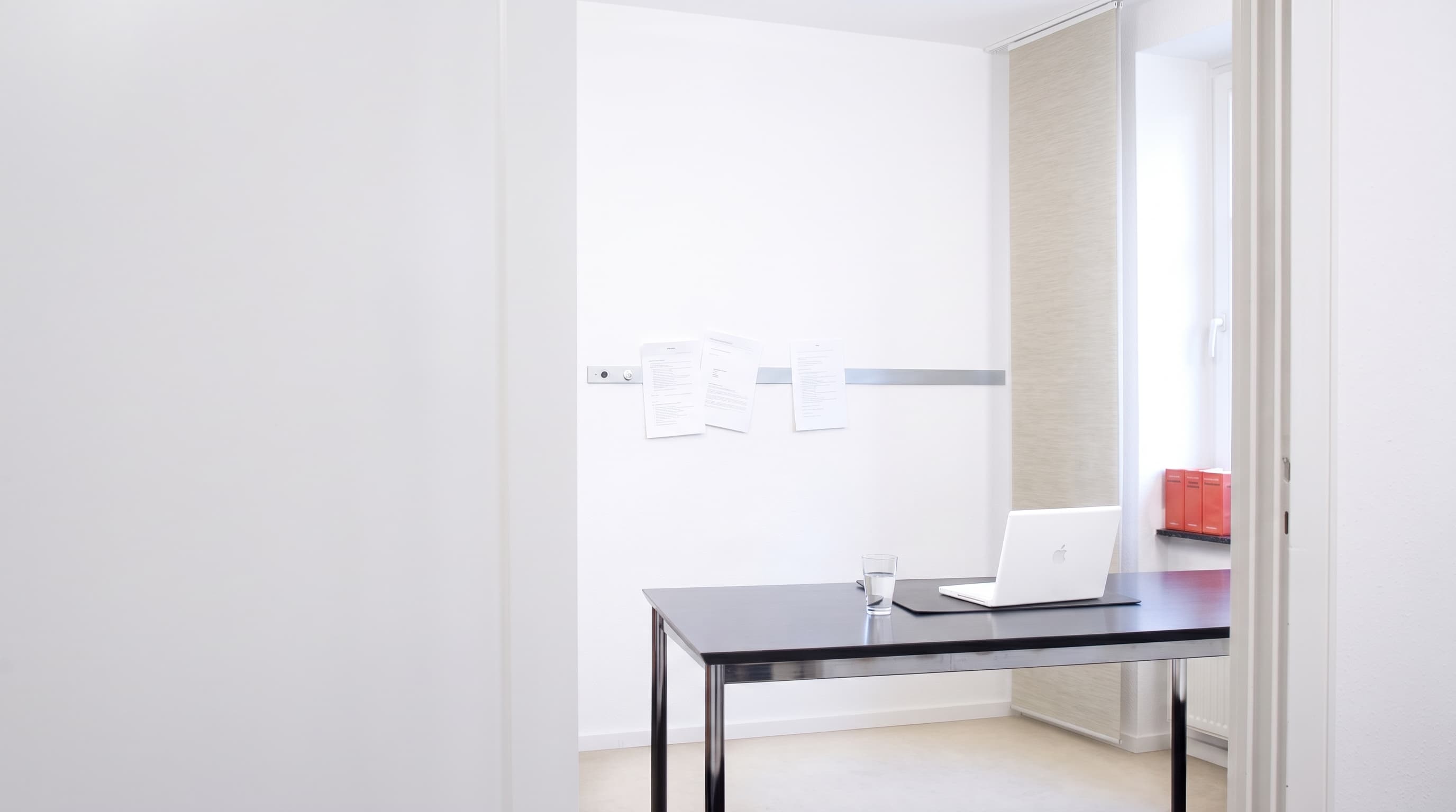 Helles Besprechungszimmer der Kanzlei mit schwarzem Tisch, Laptop, Wasserglas und roten Ordnern auf der Fensterbank